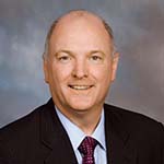 A man in a black suit, blue collared shirt, and red tie, stands and smiles